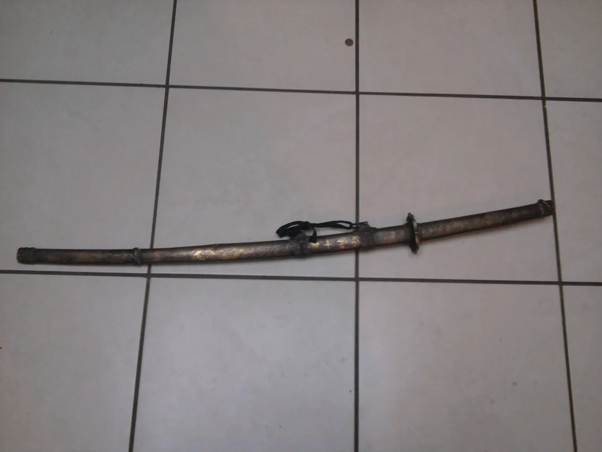 A rusty, weathered Japanese sword with a black cord tied around its hilt lies horizontally on a white tiled floor. The…