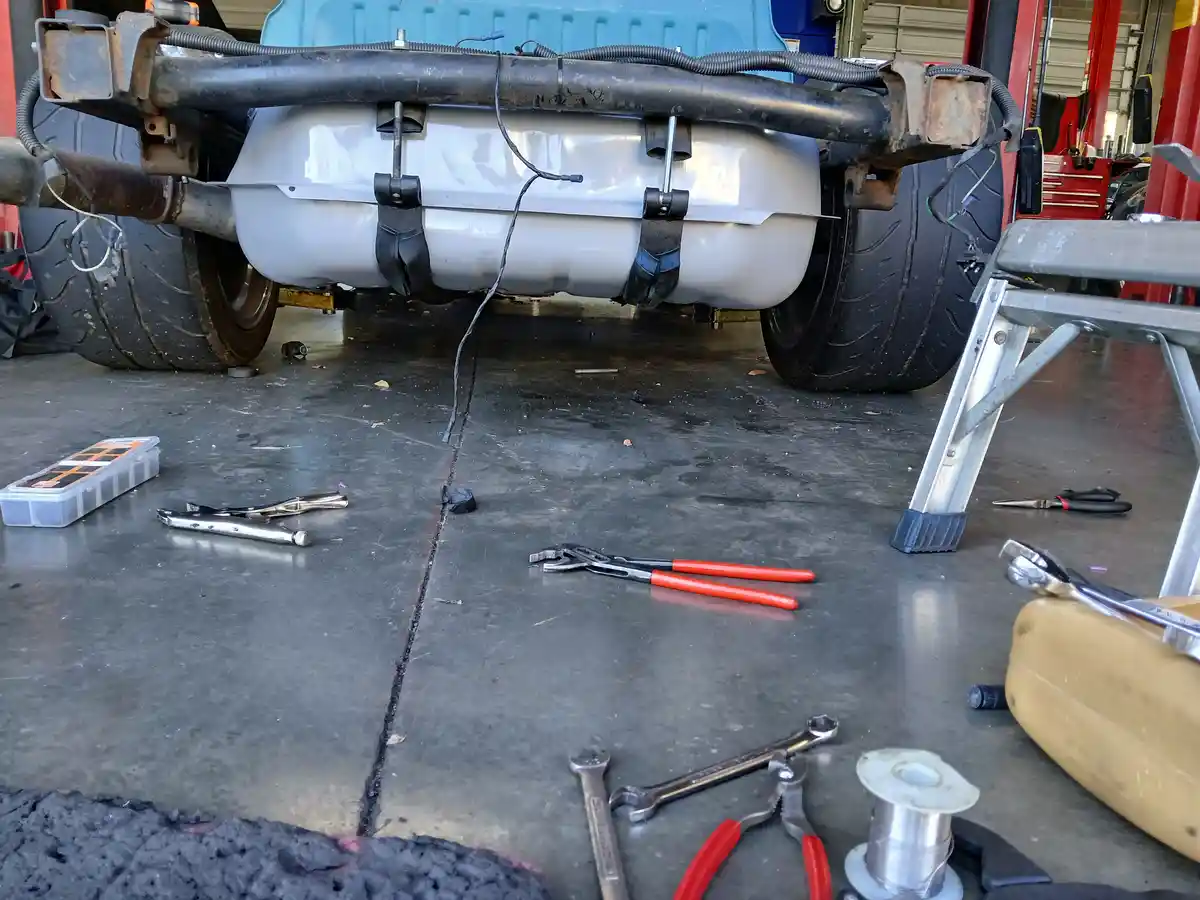 A low-angle shot of a vehicle's undercarriage in a garage, showing a silver fuel tank secured with black straps. Various…