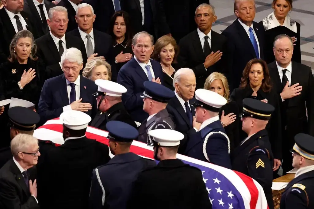 A solemn photo of U.S. presidents and their families at a state funeral, standing in formal black attire with hands over…