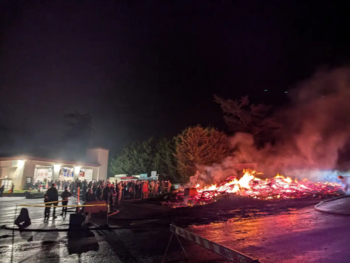 A large fire burns intensely at night in a paved area, casting red-orange light and smoke, with emergency responders and…