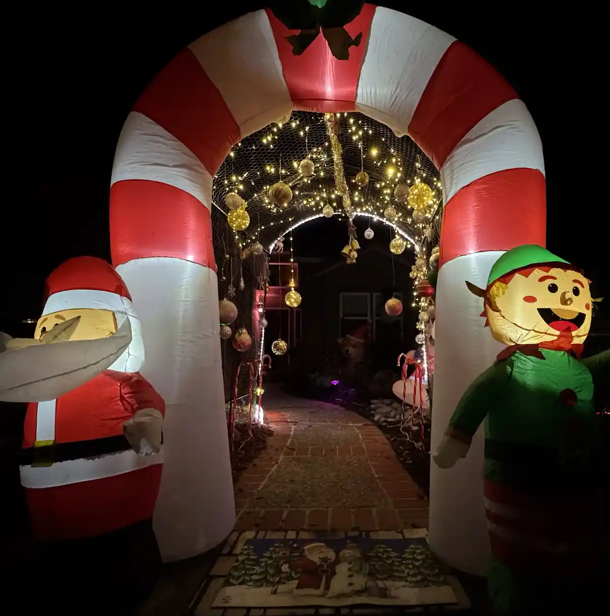 An inflatable candy cane archway flanked by a Santa and an elf, leading to a decorated walkway at night. The arch is adorned…