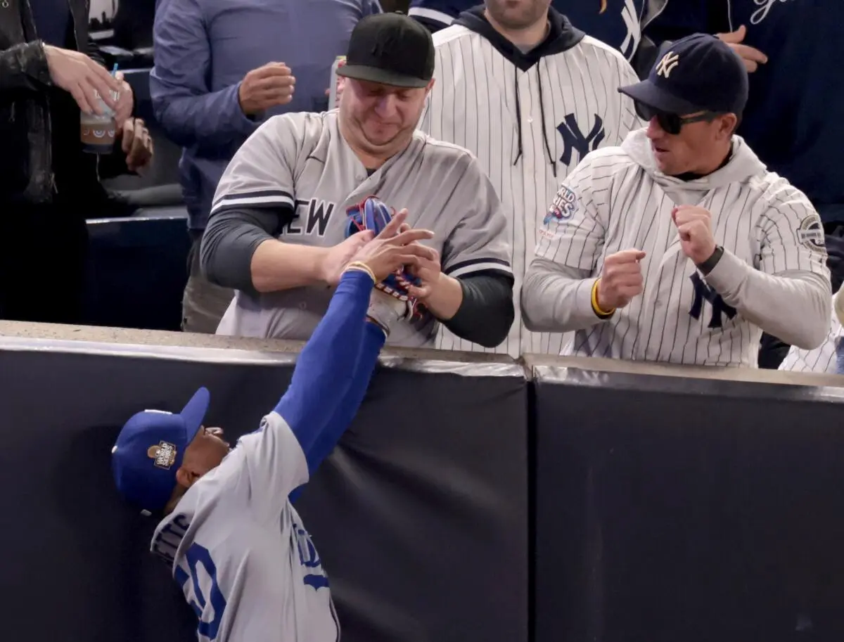 A baseball player in a blue cap and grey uniform leans over a black barrier, reaching up with his glove to high-five a…