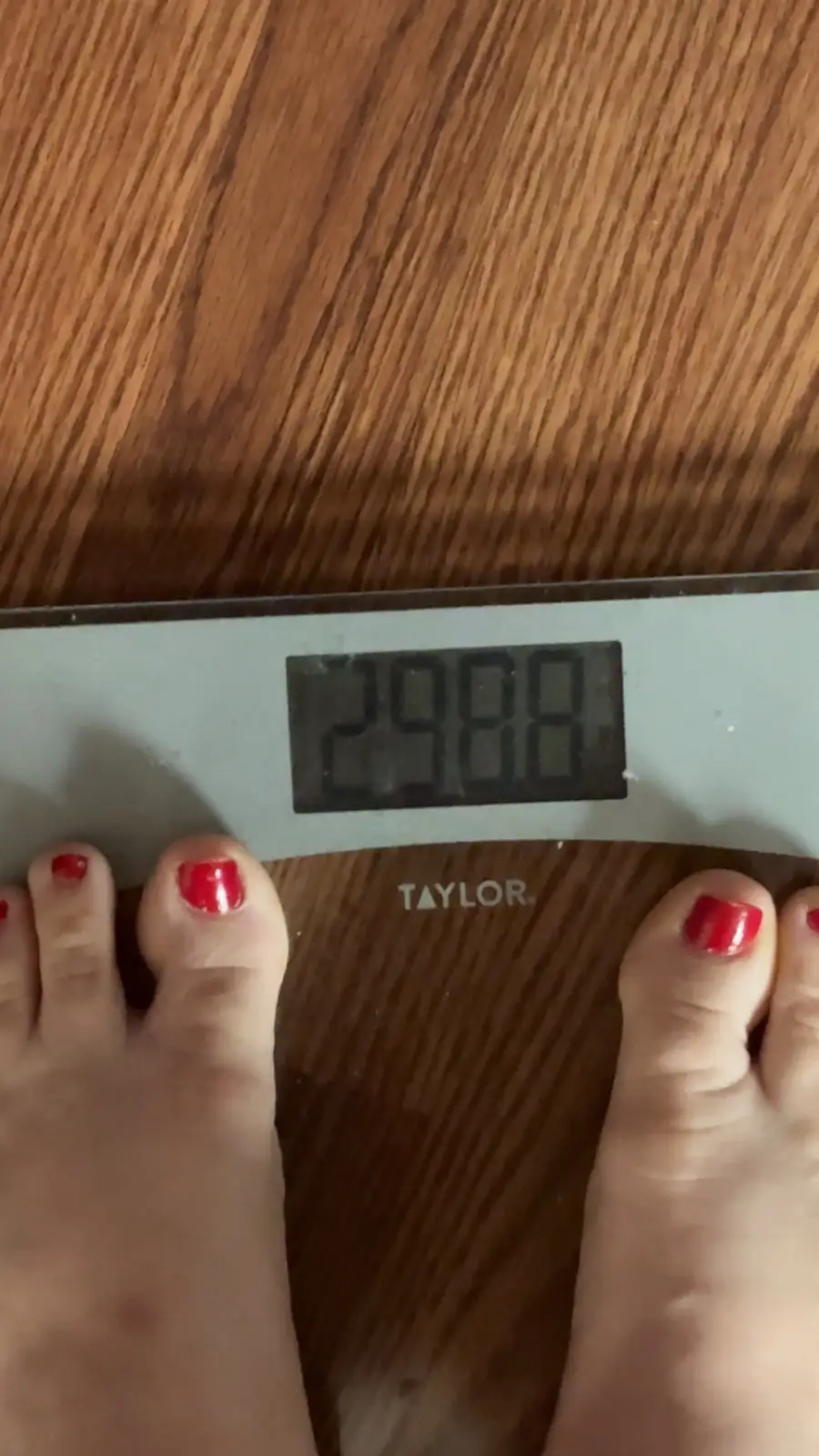 A close-up shot shows a person's feet with red-painted toenails standing on a digital bathroom scale branded 'TAYLOR'. The…