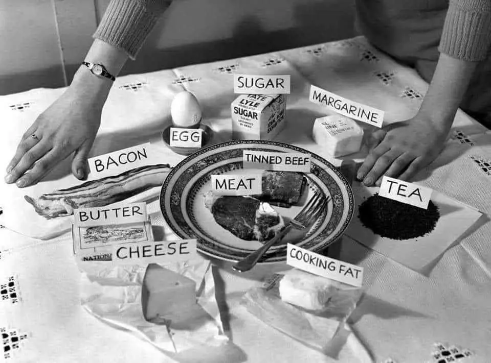 A black-and-white photo shows a person's hands resting on a table with various food items labeled with handwritten signs. On…