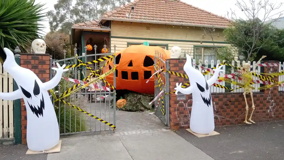 A residential front yard is heavily decorated for Halloween with an oversized inflatable pumpkin house, two inflatable…