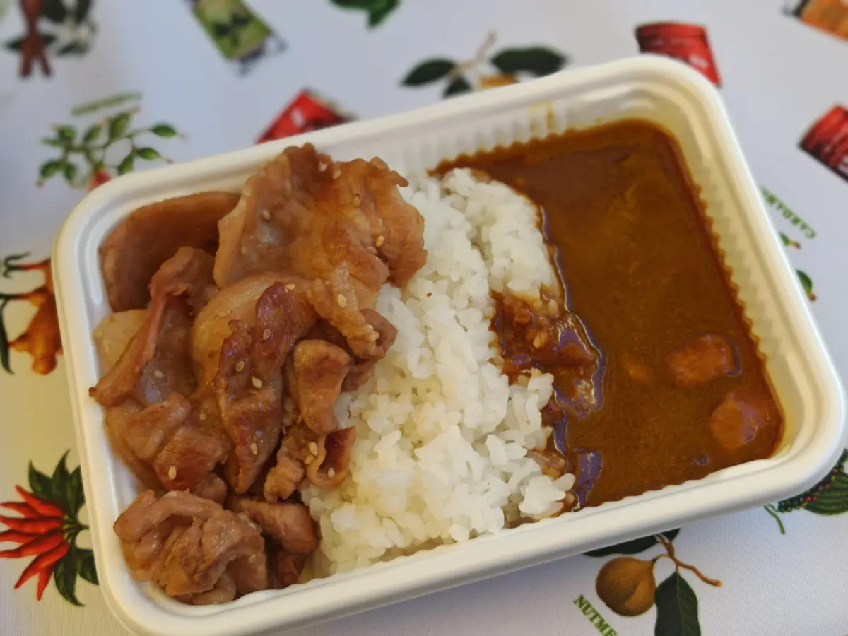 A white takeout container holds a meal with white rice, brown curry sauce, and pieces of meat with sesame seeds. The text…