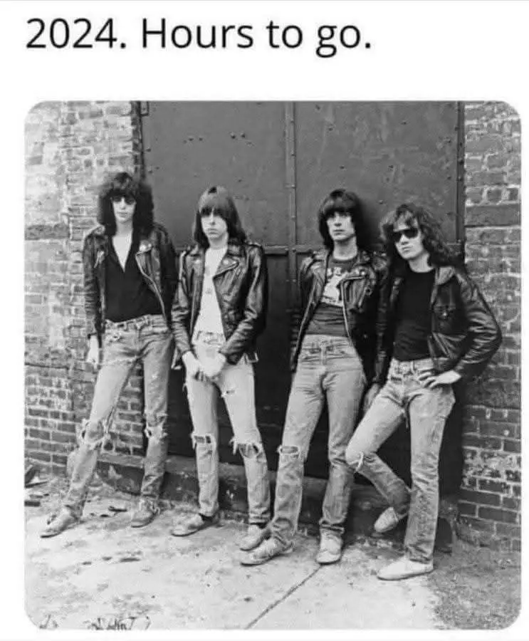 A black-and-white photo shows four men in 1980s-style leather jackets and ripped jeans leaning against a brick wall. The…