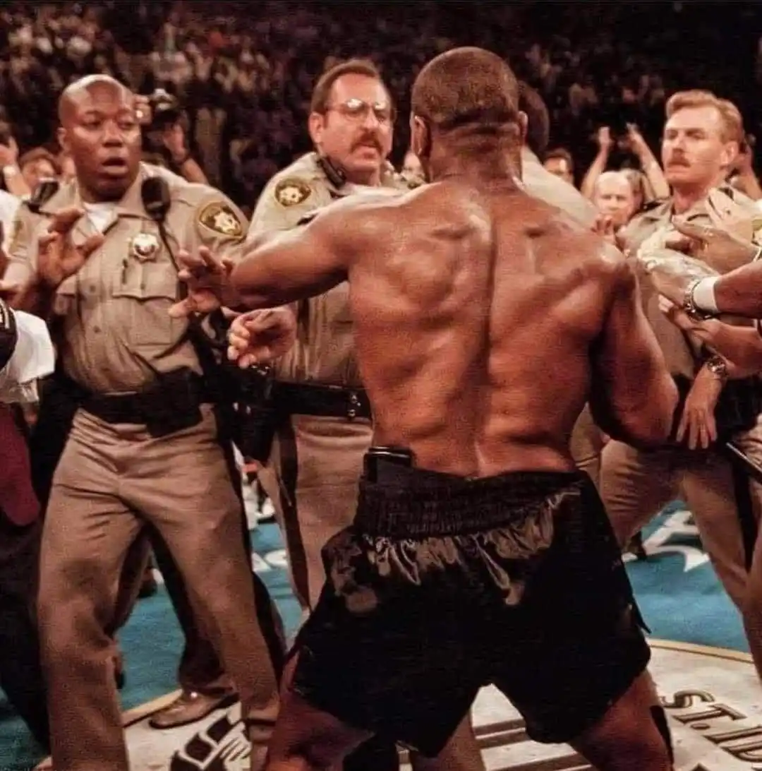 A muscular, shirtless man in black boxing shorts stands with his back to the camera, surrounded by multiple uniformed police…