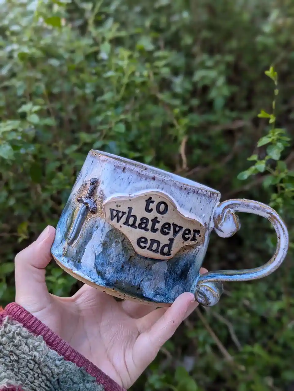 A hand holds a handmade ceramic mug with a blue and white speckled glaze, featuring a raised plaque with the text 'to…