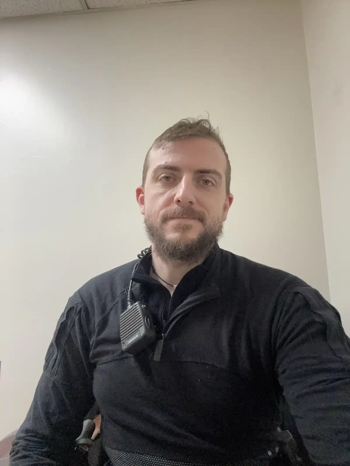 A man with a beard and short hair, wearing a black zip-up jacket, looks directly at the camera with a neutral expression. A…