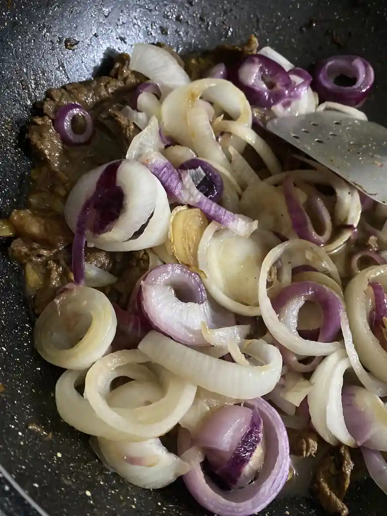 A close-up photo shows sliced red and white onions sautéing with browned meat in a dark non-stick pan, with a metal spatula…