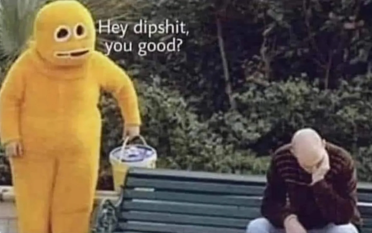 A person in a full-body yellow mascot costume with large round eyes and a simple mouth stands next to a park bench, holding…