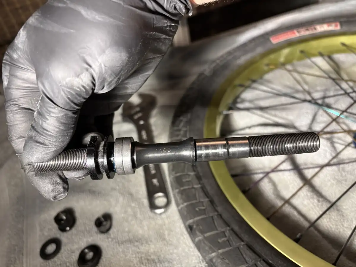 A gloved hand holds a bicycle hub axle with the number '1109' engraved on it, with a bike wheel and tools in the background…