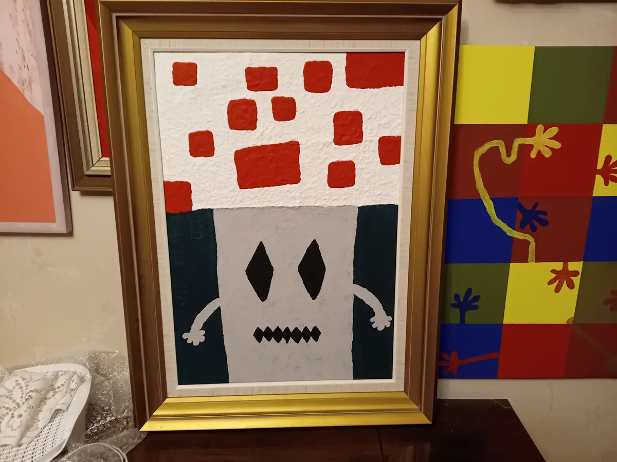 A framed painting depicts a simple, stylized monster with a white body, black diamond eyes, a jagged mouth, and two arms…