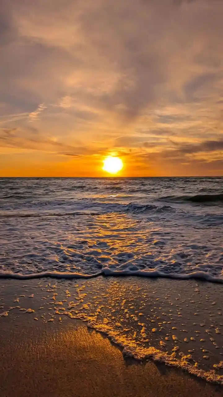 A serene beach sunset with the sun low on the horizon, casting golden light across the ocean waves and wet sand. The image…