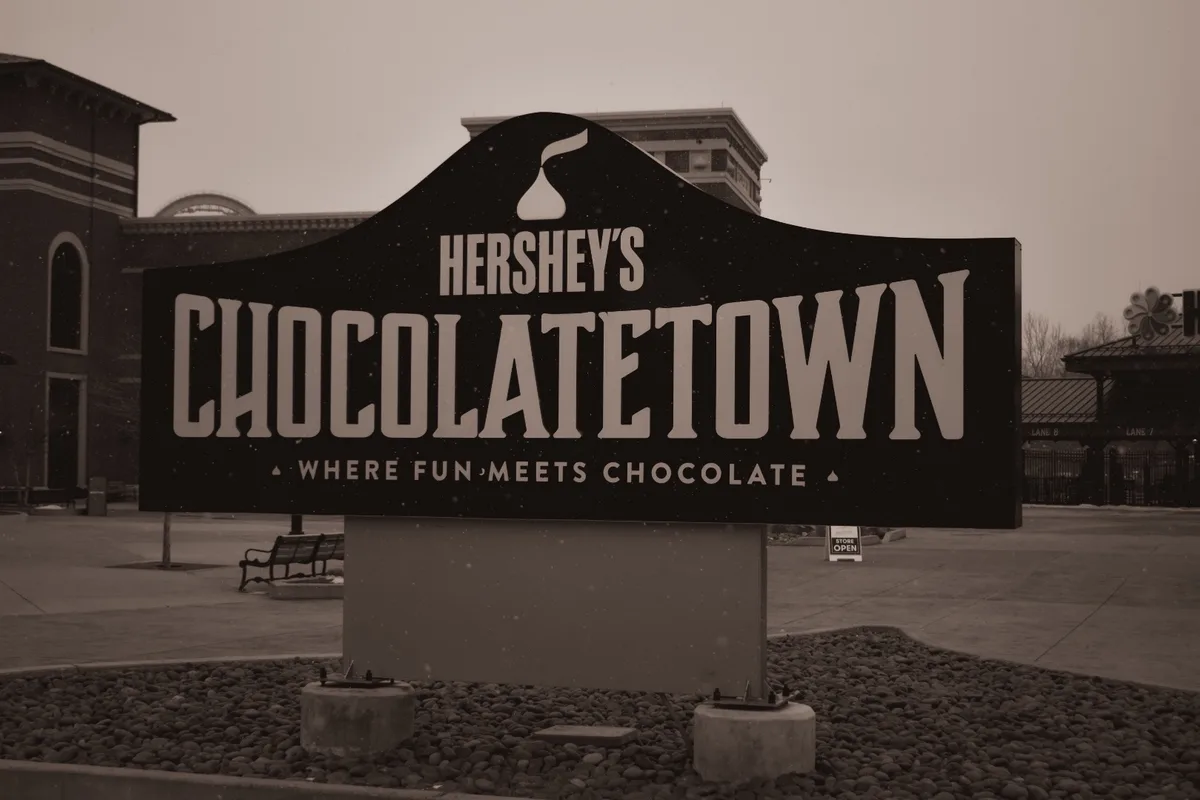 A sepia-toned photo shows the large black sign for Hershey's Chocolate Town, which reads 'WHERE FUN MEETS CHOCOLATE' beneath…
