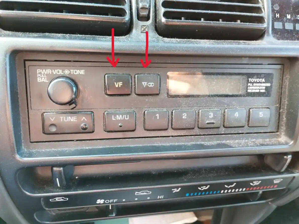 A close-up of a dusty Toyota car radio with red arrows pointing to two buttons labeled 'VF' and '7-∞'. The radio has a…