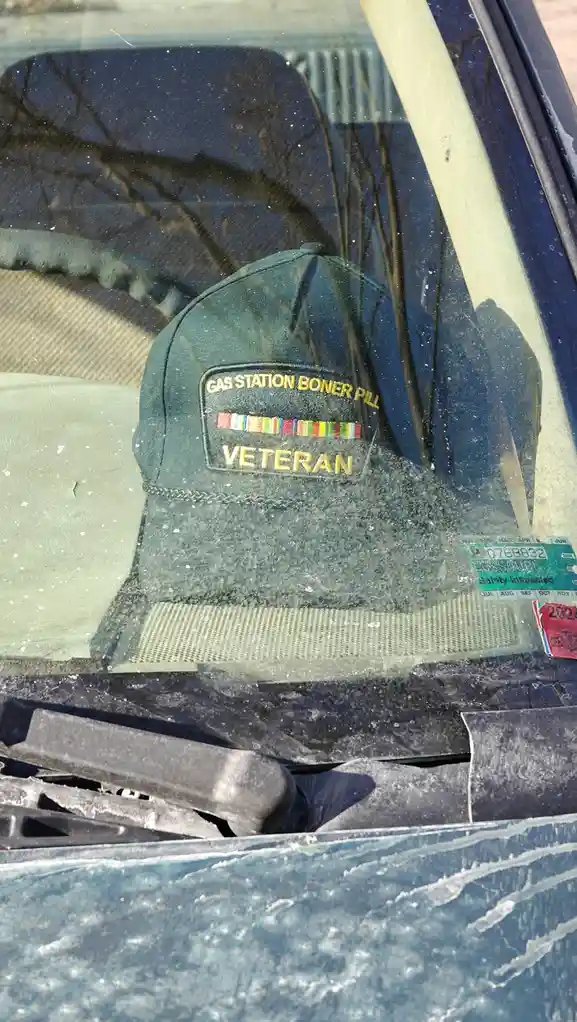 A green baseball cap with a patch reading 'GAS STATION BONER PILL' above a row of colorful ribbons and 'VETERAN' below sits…