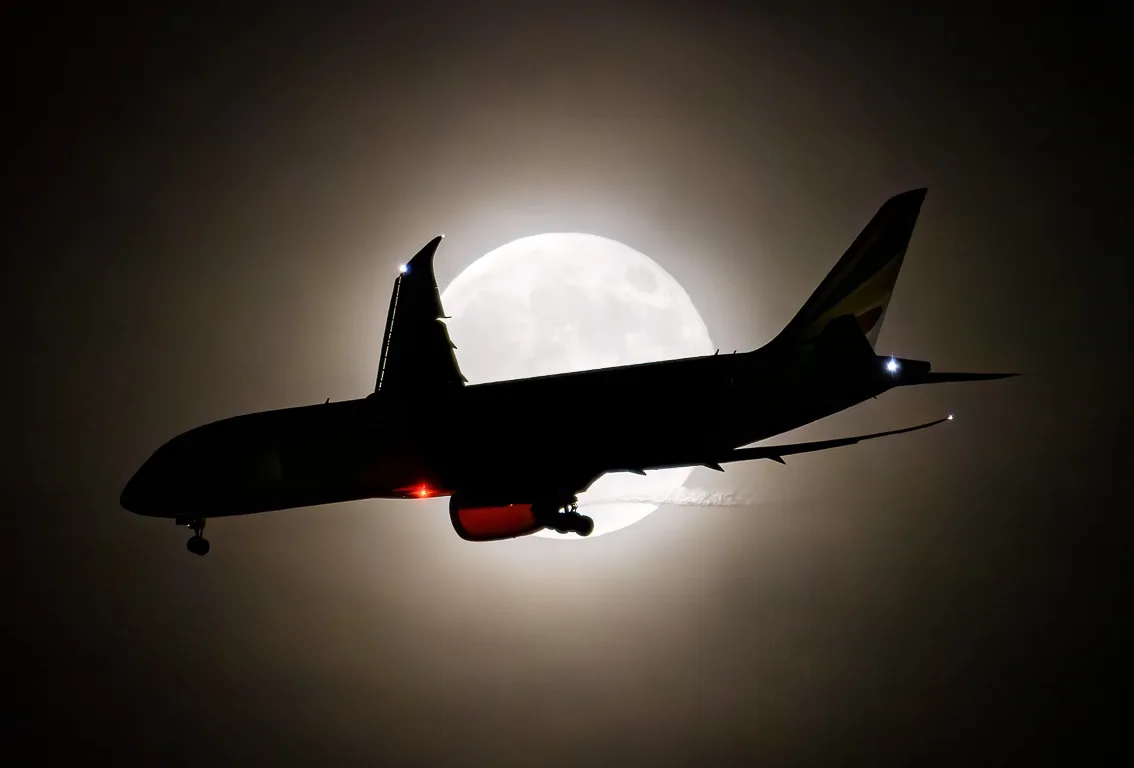 A silhouette of a commercial airplane flying against a large, bright full moon. The plane's landing lights are on, and its…