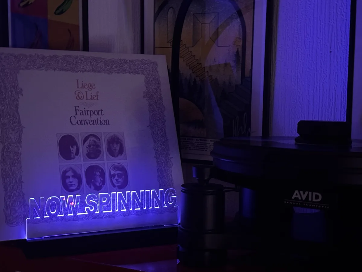 A vinyl record player, branded 'AVID SEQUOIA TURNTABLE', sits next to a glowing 'NOW SPINNING' sign. The sign displays the…