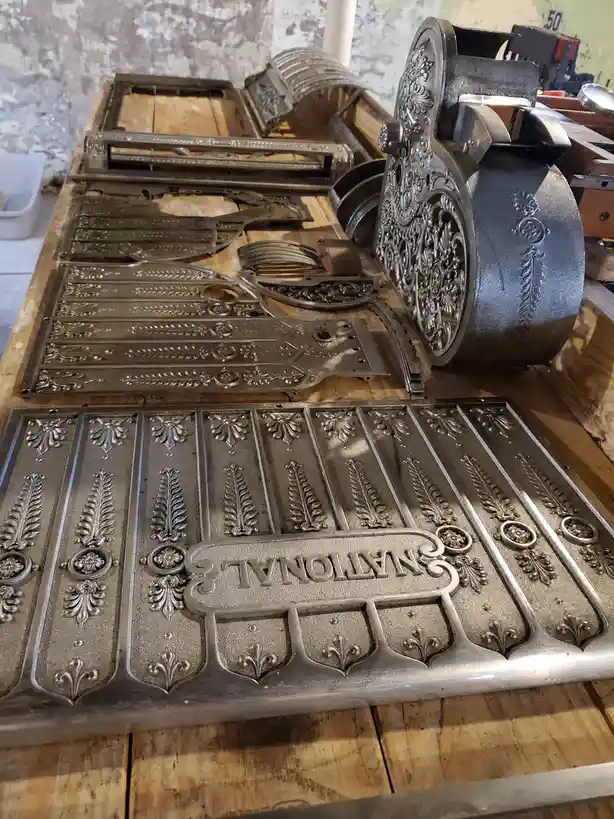 A disassembled vintage National cash register is laid out on a wooden table, showing ornate metal plates with floral and…
