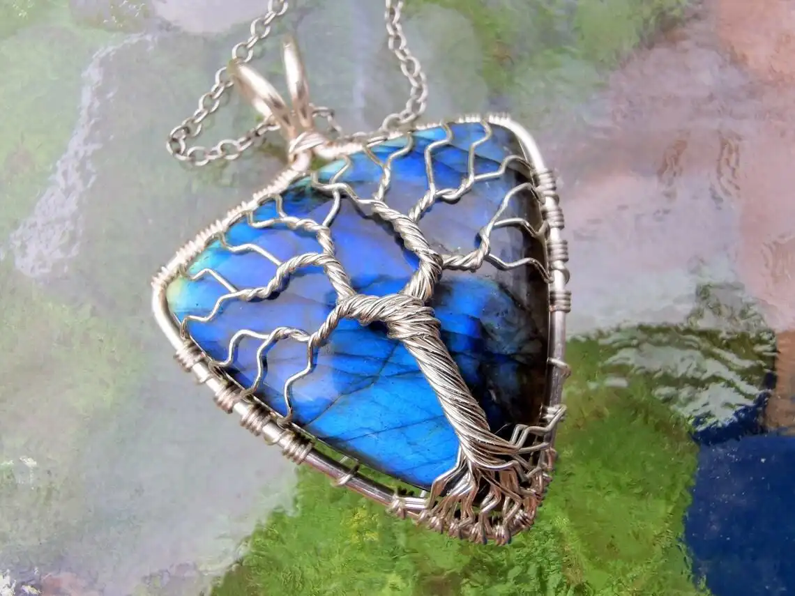 A close-up photo of a silver wire-wrapped pendant featuring a blue labradorite stone shaped like a heart, with a…