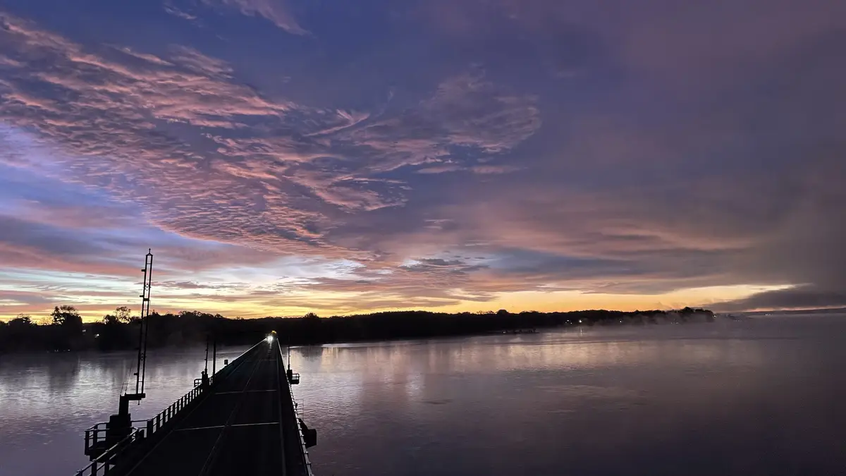 A serene photograph of a train bridge at dawn or dusk, with a train's headlight visible in the distance. The sky is painted…