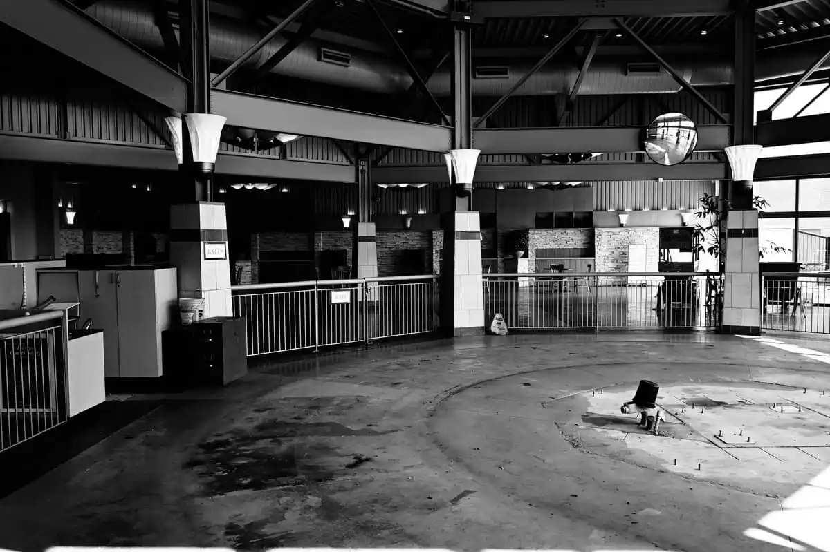 a black-and-white photo shows an empty, abandoned indoor space with exposed beams, railings, and pillars. A bucket and mop…