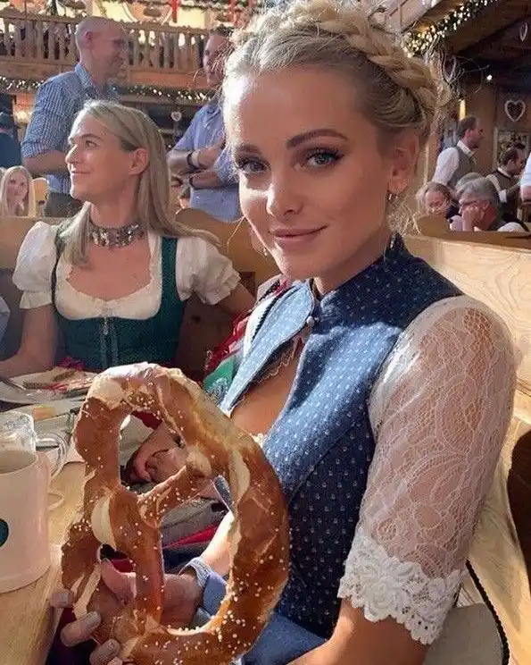 A woman in traditional German attire, including a blue vest and lace sleeves, sits at a wooden table in a rustic restaurant…