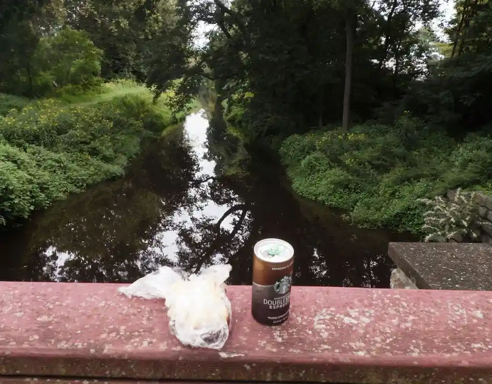 A Starbucks Doubleshot Espresso can and a plastic-wrapped food item sit on a weathered red railing overlooking a calm…