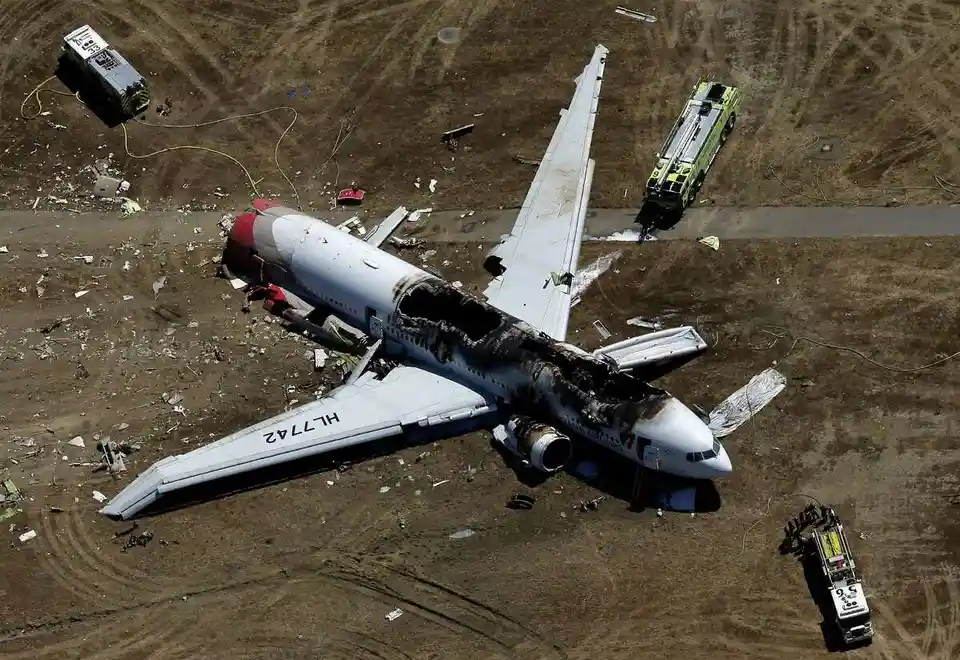 An aerial view shows a severely damaged and burnt airplane lying on a dirt field, surrounded by emergency vehicles and…