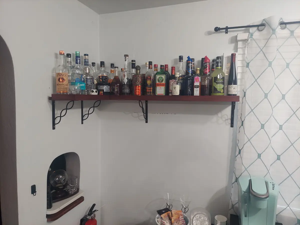 A wooden shelf mounted on a white wall in a bathroom holds numerous liquor bottles, including Tito's, Jägermeister, and…