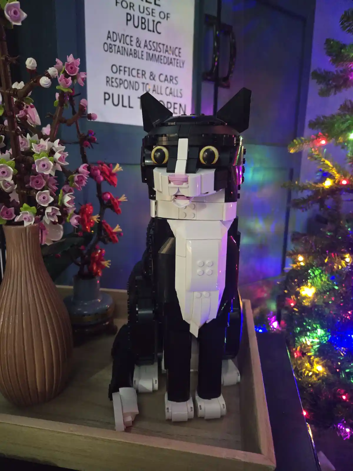 A black-and-white LEGO cat figurine sits on a wooden shelf next to a vase of pink and red flowers and a lit Christmas tree…