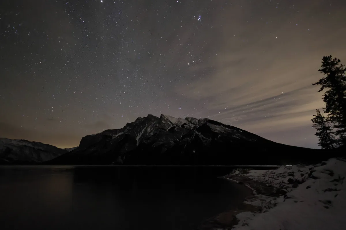 A night photograph shows a star-filled sky above a dark mountain range and a calm lake, with snow on the foreground shore…