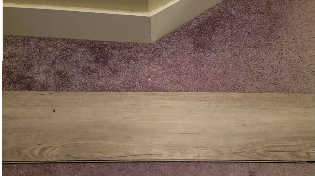 A close-up photo shows a single light wood-look laminate floor plank laid horizontally across a purple carpet, with a white…