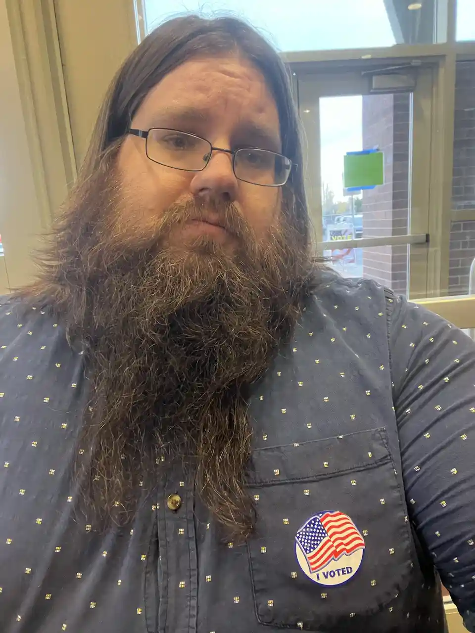 A man with long hair, a full beard, and glasses wears a dark blue patterned shirt with an 'I VOTED' sticker featuring the…