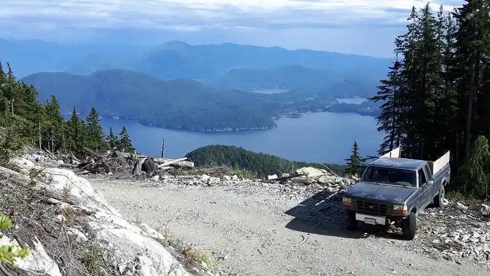 A weathered gray pickup truck is parked on a gravel overlook with a stunning panoramic view of a vast blue lake surrounded…
