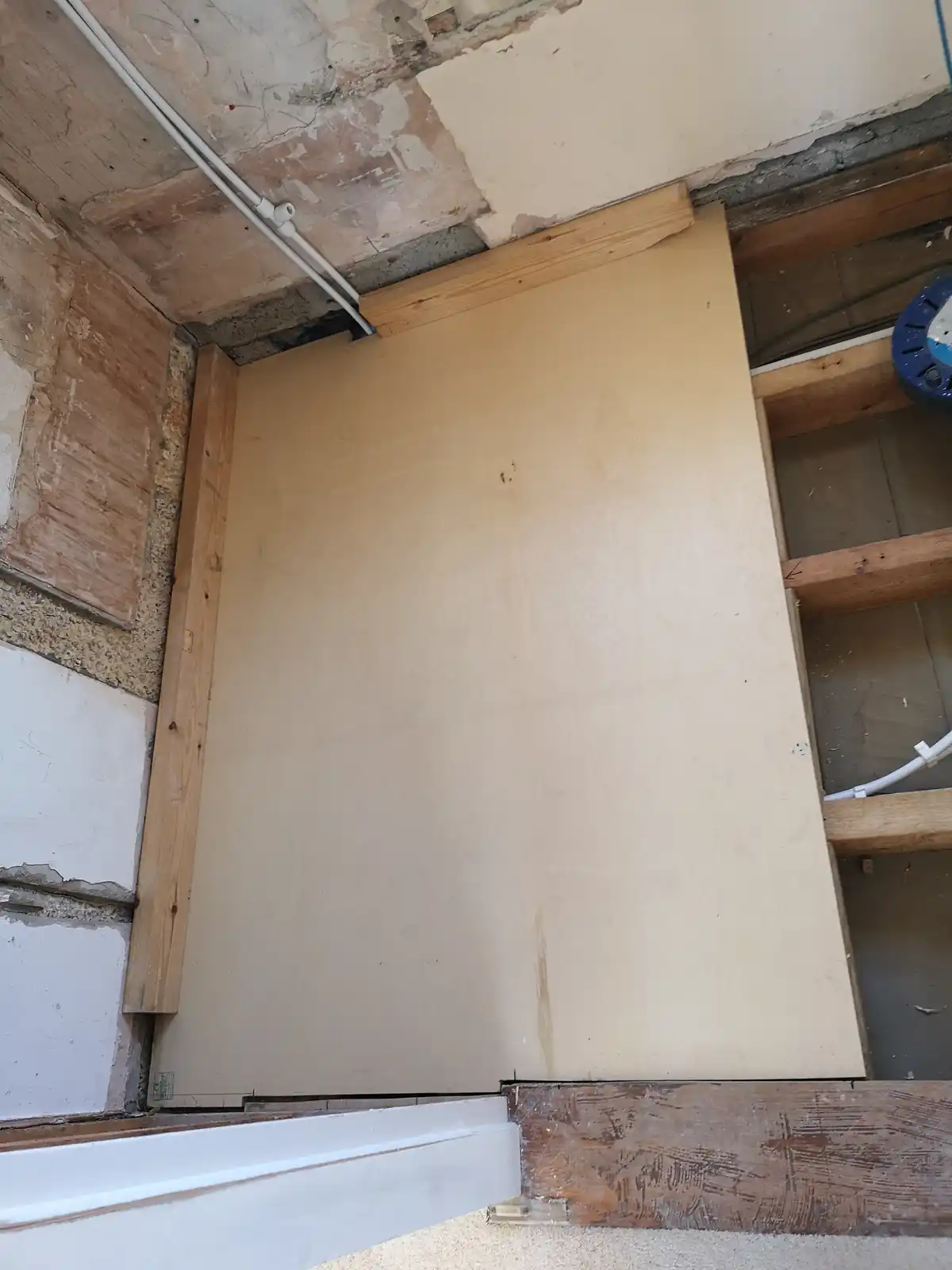 A large, plain plywood sheet is framed with wooden beams and installed in a doorway, replacing a wall with visible exposed…