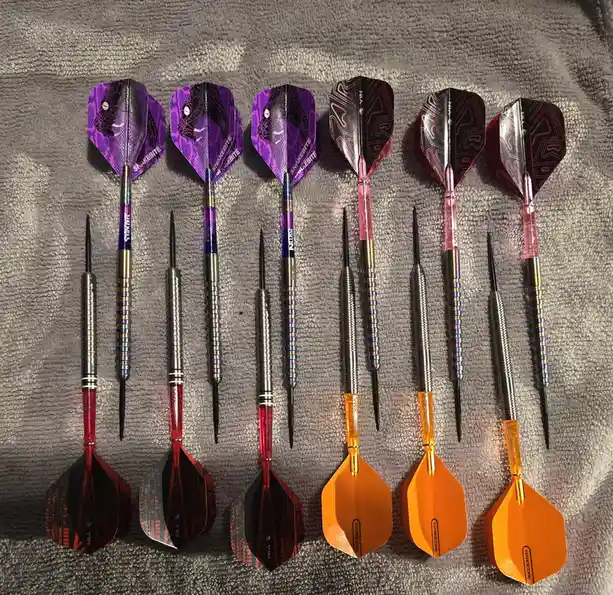 Six darts are arranged in two rows on a gray towel, each with distinct colorful flights and shafts. The top row has purple…