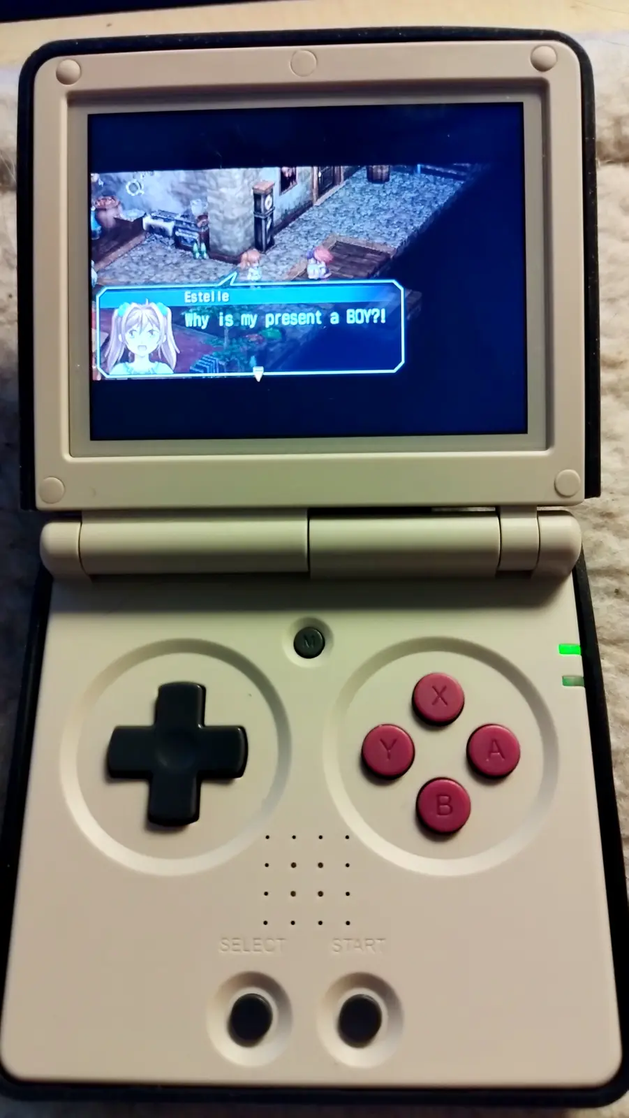 A beige handheld game console displays a scene from the game 'Tales of Vesperia' where the character Estelle exclaims, 'Why…