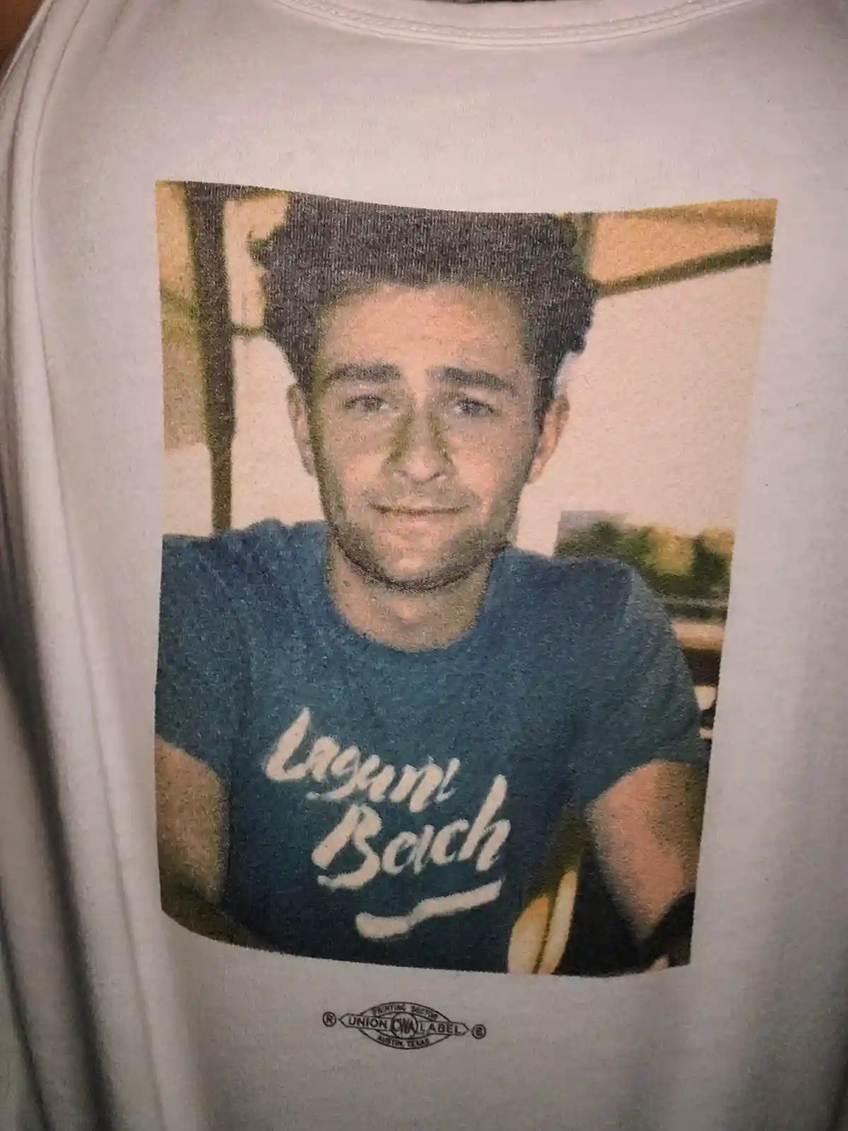 A close-up of a white t-shirt featuring a printed photo of a young man with dark hair and a slight smile, wearing a blue…