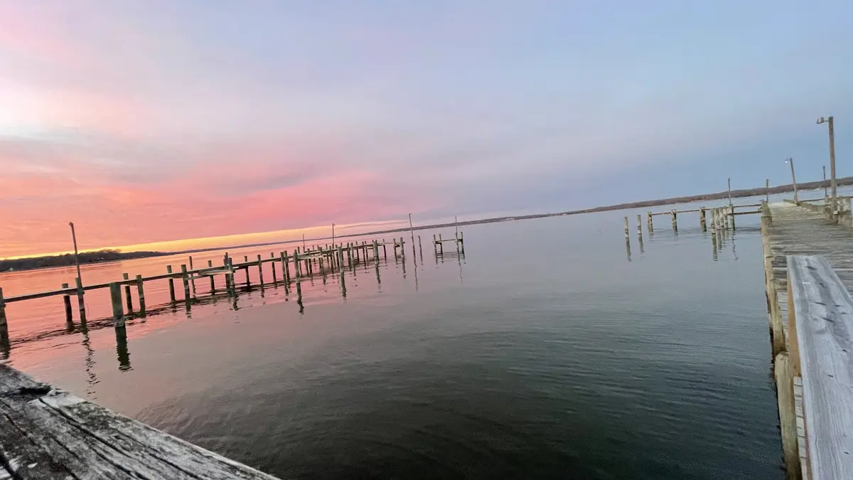 A serene photo of a wooden pier extending into calm water during a colorful sunset with pink and orange hues in the sky. The…