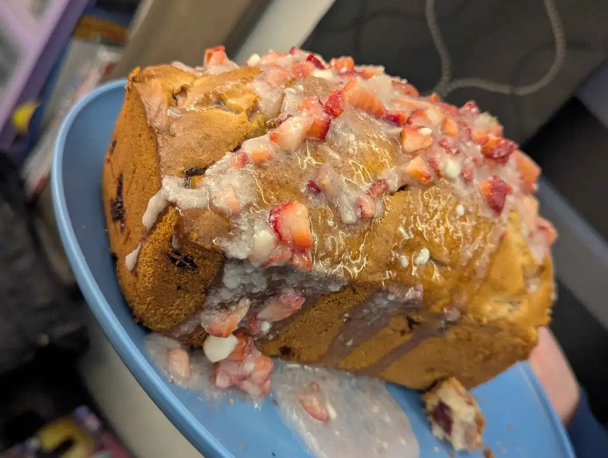 A close-up photo shows a homemade loaf cake on a blue plate, covered in white icing and topped with chopped red…