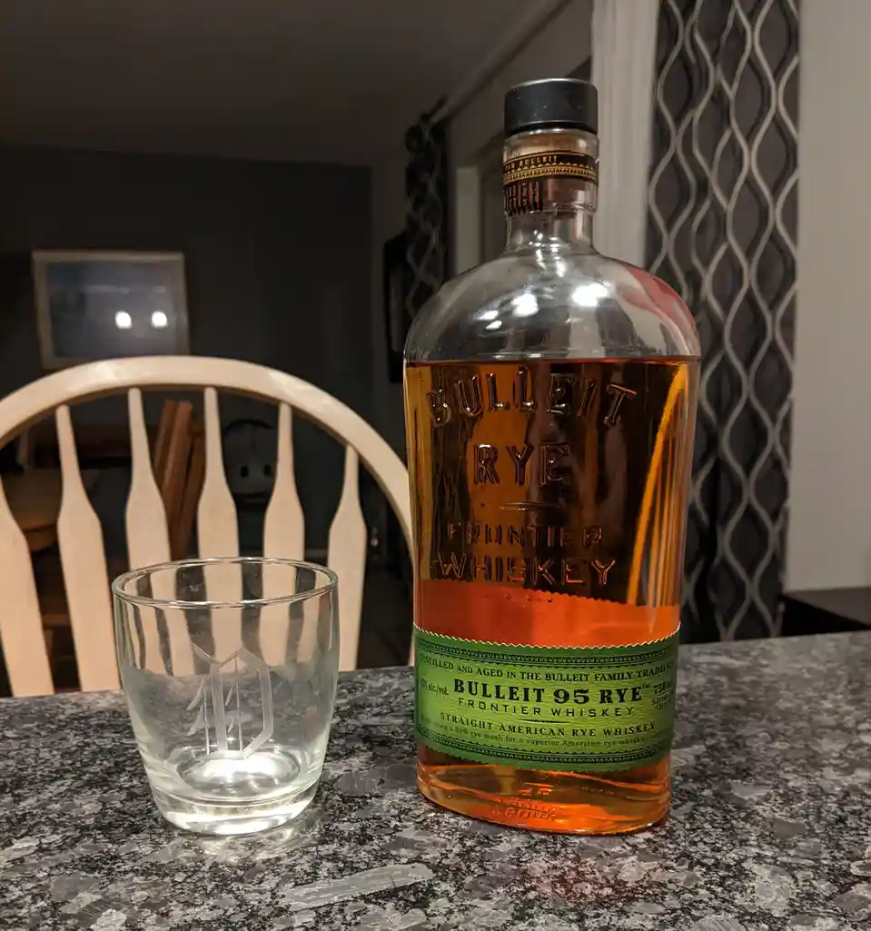 A bottle of Bulleit 95 Rye Frontier Whiskey sits next to an empty glass on a granite countertop, with a wooden chair and…