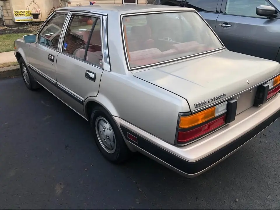 A silver 1980s Nissan Stanza sedan is parked outdoors, viewed from the rear three-quarter angle. The car has a beige…