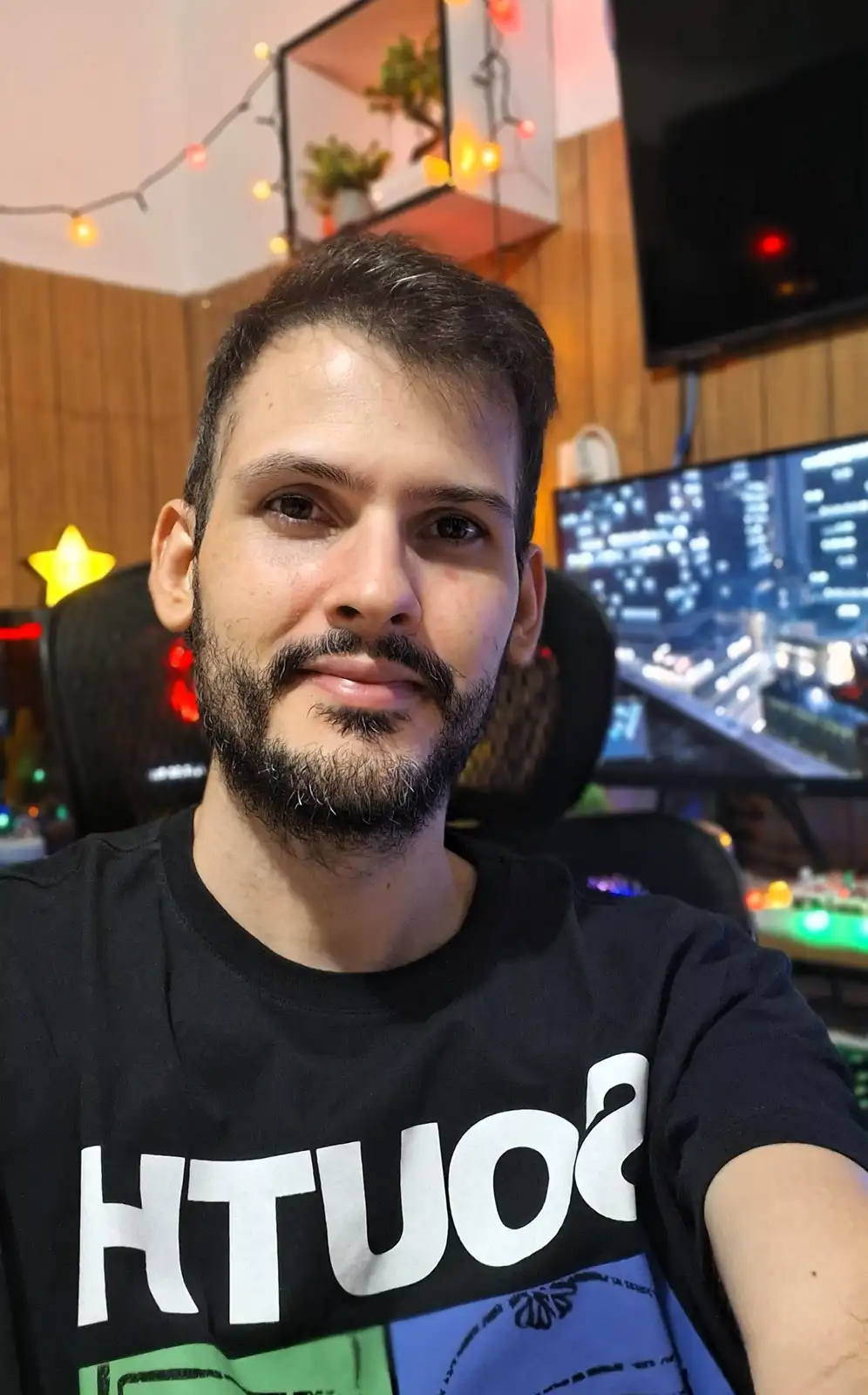 A man with a beard and mustache takes a selfie in a room with string lights, a gaming chair, and multiple monitors…