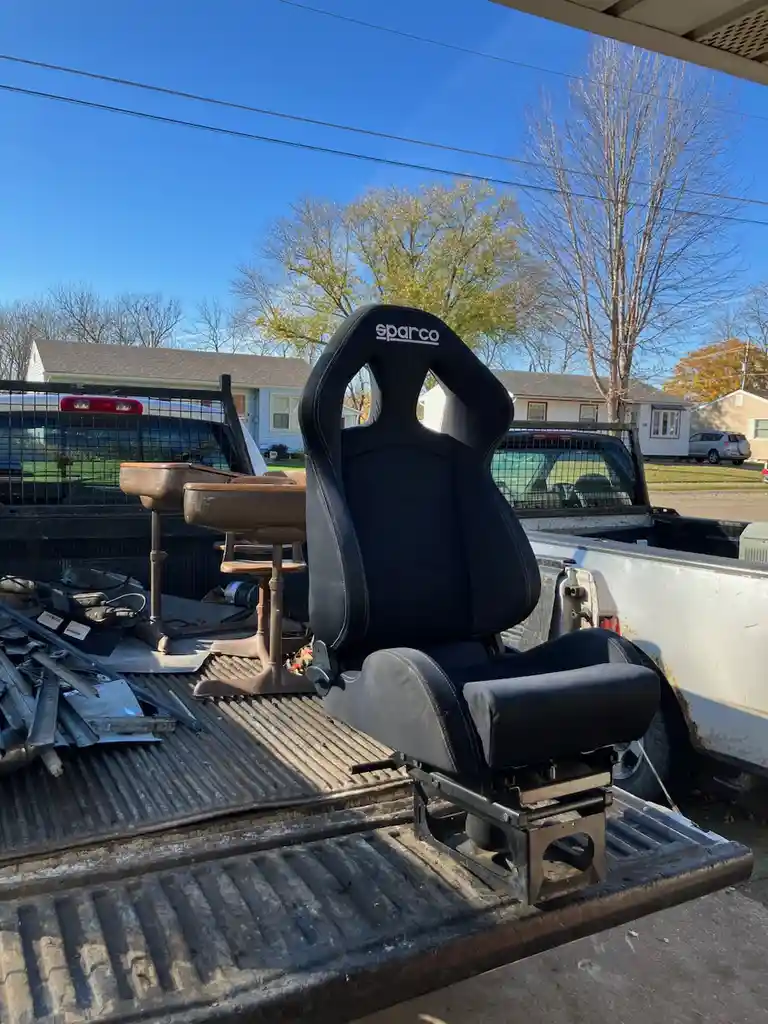 A black Sparco racing bucket seat sits prominently in the bed of a pickup truck, surrounded by clutter including old wooden…
