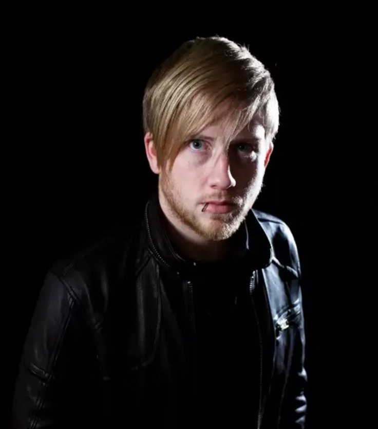 A man with blond hair and a beard, wearing a black leather jacket, stares intensely at the camera against a black…
