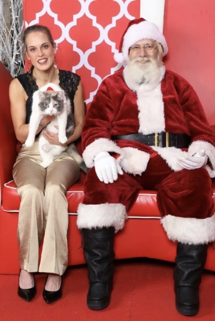 A woman in a black top and beige pants sits on a red couch holding a gray and white cat, next to a man dressed as Santa…