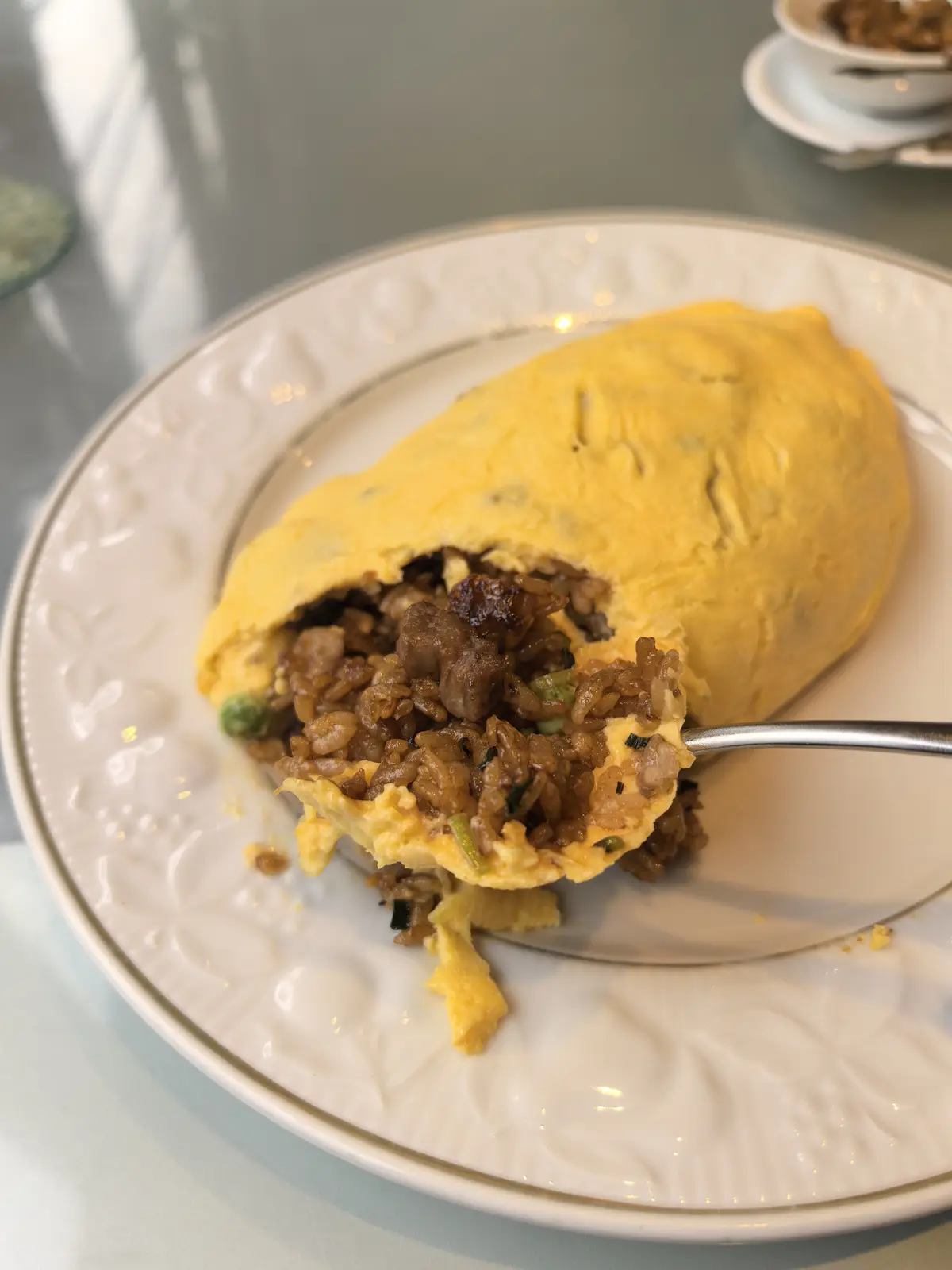 A close-up photo shows a golden-yellow omelette on a white plate with a fork lifting a bite, revealing fried rice and meat…